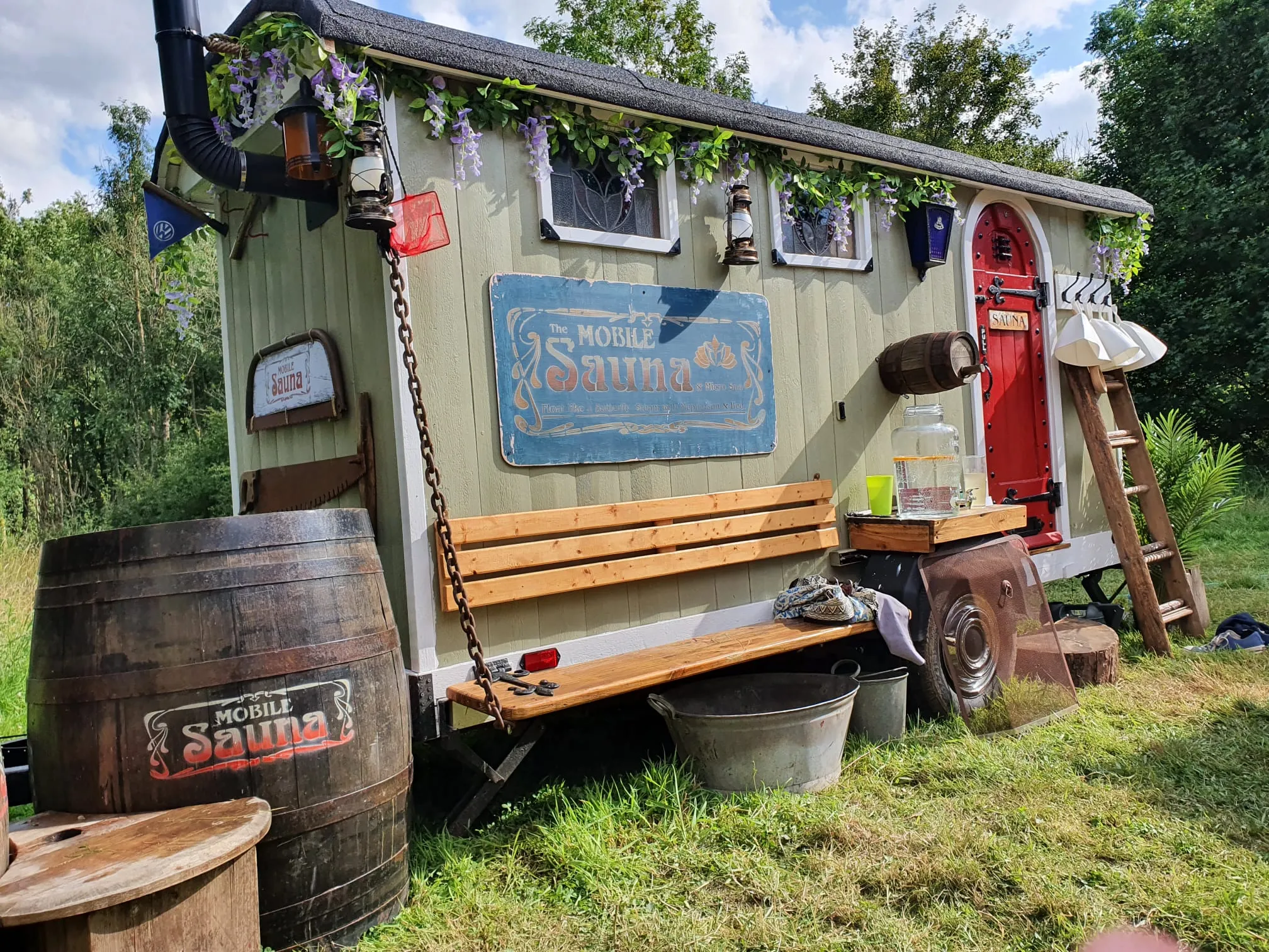 The sage-green shepherd’s hut sauna with its red arched door, parked in an open meadow